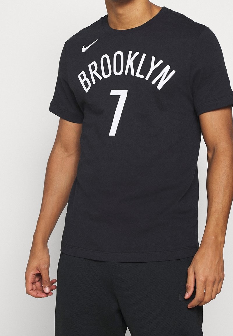 Homme portant un t-shirt noir Nike avec "BROOKLYN 7" imprimé en blanc sur le devant, associé à un pantalon noir.