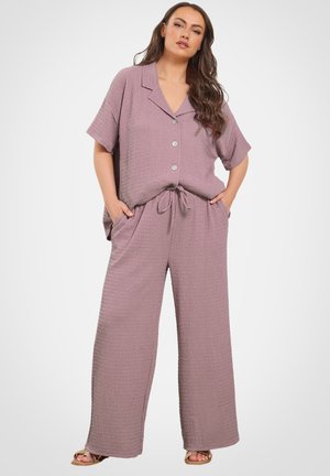 Vrouw staand met handen in de zakken, gekleed in een mauve tweedelige outfit met korte mouwen en wijde broekspijpen.