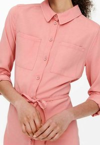 Camicia button-up rosa con colletto, due tasche sul petto e un nastro in vita. Tessuto liscio con maniche lunghe e dettagli a bottoni visibili.