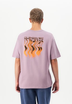 Mann iført en lys lilla t-skjorte med et forvrengt oransje og svart grafisk design på baksiden, kombinert med blå bukser.