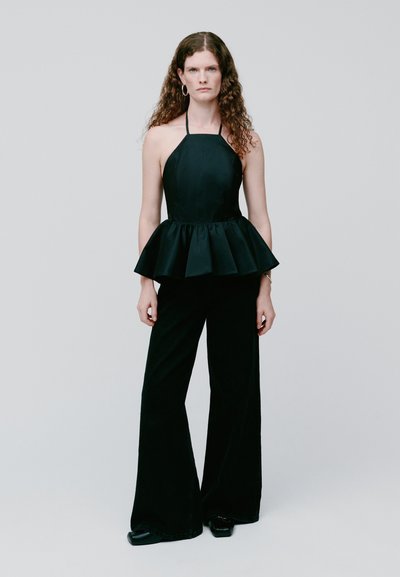 Femme aux longs cheveux bouclés porte un top dos nu noir avec une taille peplum et un pantalon noir à jambes larges, debout devant un fond clair uni.