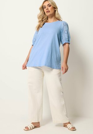 Donna che indossa una maglietta azzurro chiaro a maniche corte a sbuffo con dettagli ad occhiello, pantaloni bianchi a gamba larga e sandali bianchi slip-on, in piedi davanti a uno sfondo semplice.