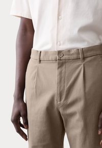 Pantalones beige con textura, cierre de botón, pinzas y cintura con trabillas, combinados con una camisa blanca de manga corta.