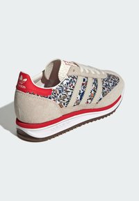 Beige mockasin-sneakers med blomstrande tygöverdel, röda detaljer på klacken och sulan, tre randig design, och vita snören. Texturerad gummiyttersula.