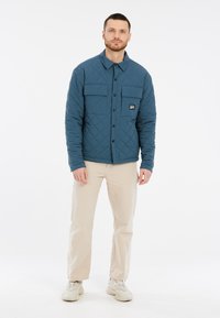 Gequiltede blauwe jas met twee borstzakken en zwarte hardware, gecombineerd met beige broeken. Het ontwerp heeft een kraag en een knoopsluiting aan de voorkant.