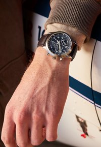 Montre chronographe avec un cadran noir, des marquages blancs et un bracelet en cuir brun. Boîtier en acier inoxydable avec trois sous-cadrans et des poussoirs.