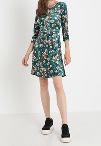 Grünes Kleid mit Blumenmuster, Dreiviertelärmel, tailliertem Schnitt und ausgestellt Rock. Kombiniert mit schwarzen Sportsneakern.