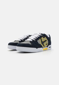 Navy blue suede sneakers with white laces, gray accents, and a yellow logo design; rubber sole with textured grip for stability.