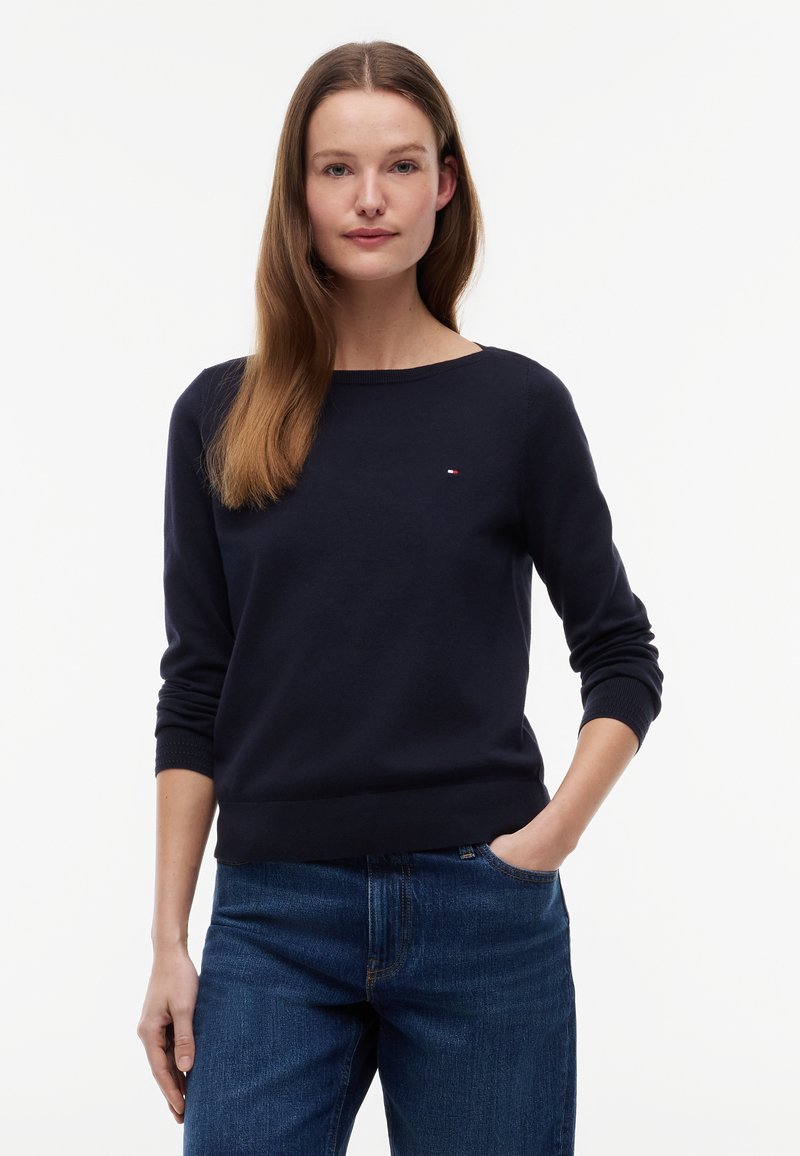 Donna con lunghi capelli castani che indossa un maglione blu navy e jeans blu, in piedi con una mano in tasca contro uno sfondo bianco.