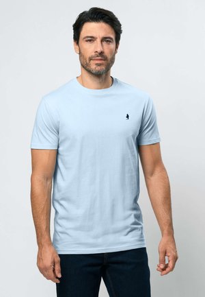 Mand med kort mørkt hår og skæg iført lyseblå kortærmet t-shirt og mørke bukser, står foran en ensfarvet lys baggrund.