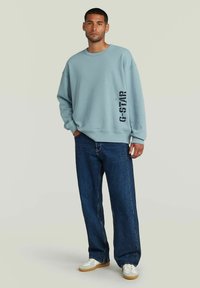 Sudadera azul claro de corte relajado con puños acanalados, que presenta un logo negro impreso en la manga izquierda. Combinada con unos vaqueros oscuros holgados y zapatillas blancas.