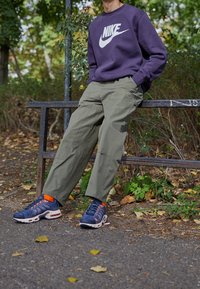 Nike purple sweatshirt with white logo, olive green cargo pants, and navy sneakers with orange accents. Set against a natural background.