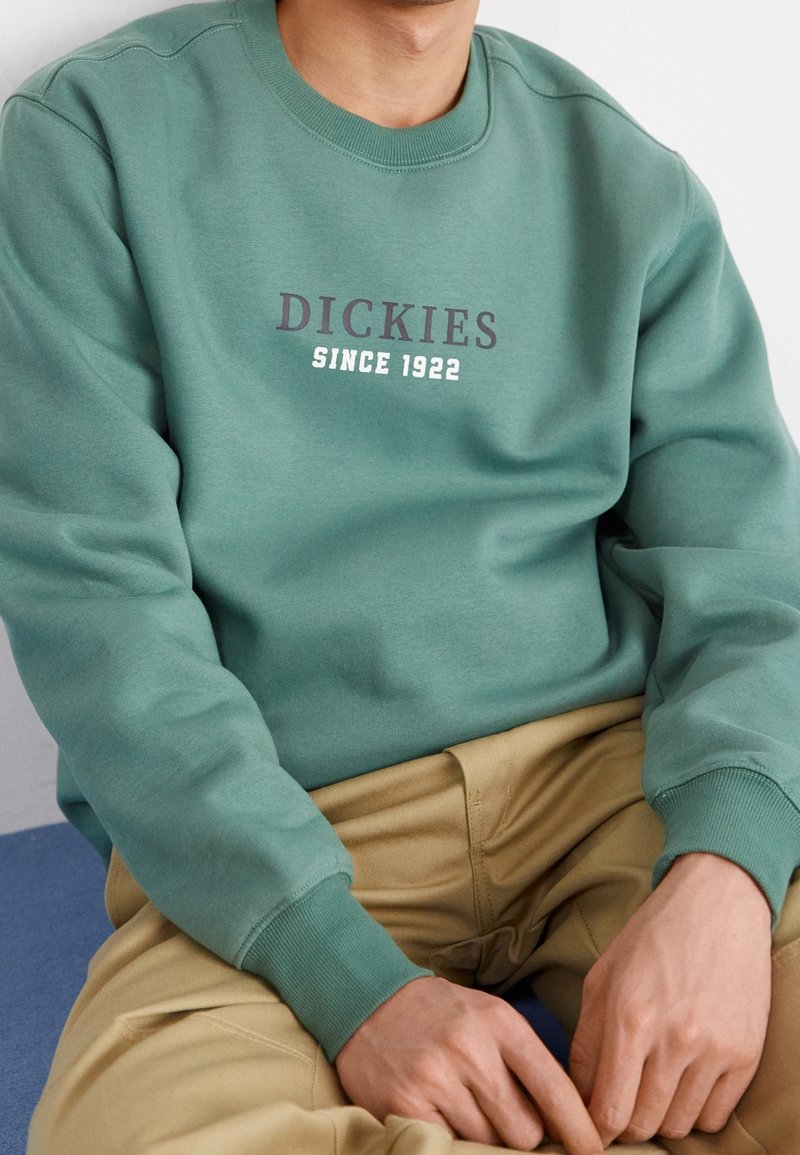 Man i grön tröja med texten "DICKIES SINCE 1922", beige byxor, händerna vilande på knäna, sittande mot en vit vägg.