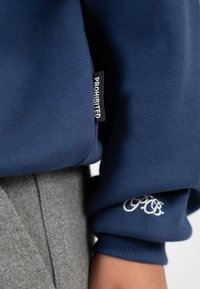 Sudadera de color azul marino con puños acanalados, que presenta una etiqueta de "PROHIBIDO" y un detalle de logo bordado en blanco. Fondo de tela gris.