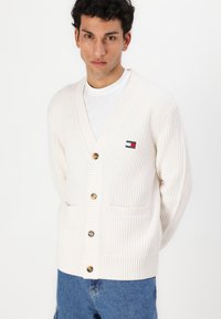 Fløyelsmyk, ribbestrikket cardigan i kremfarge med V-hals, fire knapper, to lommer og et lite logo-patch på venstre bryst.