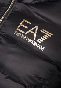 Svart vadderad jacka med glänsande yta, dragkedja och en guldfärgad logotyp med "EA" ovanför "EMPORIO ARMANI" på bröstområdet.