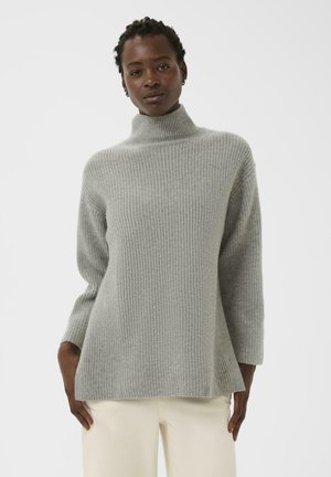 Frau mit kurzen Twists, die einen hellgrauen gerippten Rollkragenpullover und off-white Hosen vor einem schlichten hellen Hintergrund trägt.