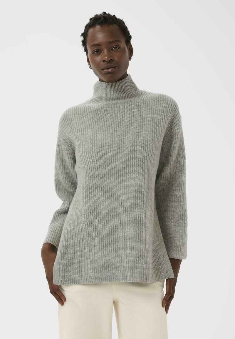 Femme avec des twists courts portant un pull col roulé côtelé gris clair et un pantalon blanc cassé sur un fond uni clair.