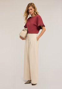 Haut à manches courtes bordeaux avec des accents plissés ; pantalon large beige ; sac à main neutre ; chaussures à bout pointu noires et brunes avec un motif serpent.