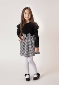 Czarny top z długim rękawem z falbankami, zestawiony z czarno-białą sukienką w pepitkę z dużą kokardą w talii. Białe rajstopy i czarne buty.