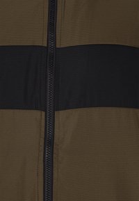 Vėjinė striukė su tamsiai žalia ir juoda spalvų bloko dizainu, tekstūruota paviršiumi, priekiniais užtrauktukais ir sustiprintomis siūlėmis.