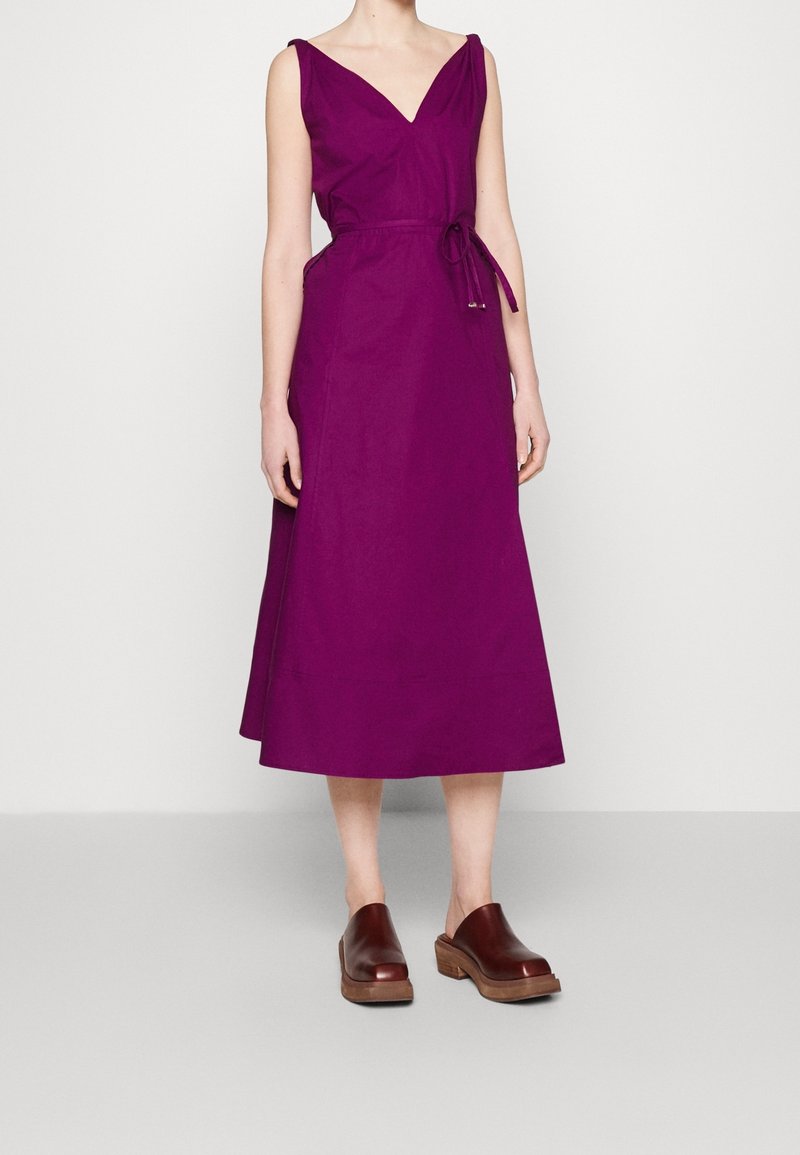 Femme portant une robe midi sans manches violette avec un col en V et une taille nouée, associée à des chaussures en cuir marron à enfiler sur un fond uni.