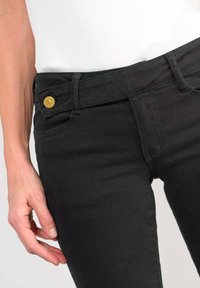 Jean en denim noir avec une coupe slim, un bouton doré à l'avant et une finition en tissu texturée. La taille se positionne au-dessus des hanches.