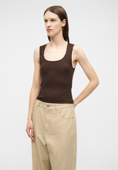 Νεαρή γυναίκα που φοράει καφέ ριμπ τοπ χωρίς μανίκια με λευκή δαντέλα και μπεζ παντελόνι φαρδύ με μικρό πουά μοτίβο, στέκεται μπροστά σε απλό φόντο.