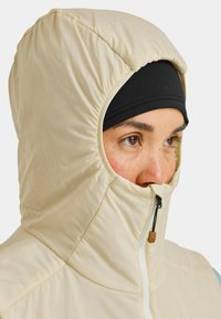 Beige isolierte Kapuze mit einer zusammengezogenen Öffnung, die eine schwarze Basislage darunter und einen Reißverschluss hat, der teilweise das Gesicht bedeckt.