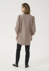 Cămașă lungă, lejeră, din țesătură taupe, cu tiv rotunjit și mâneci bufante. Poartă cu blugi negri skinny și pantofi fără toc.