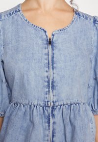 Närbild av en ljusblå denimklänning med dragkedja framtill och indraget midja, bärs av en person med delvis synlig halslinje och axlar.