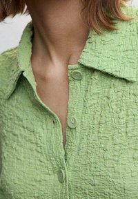 Chemise vert clair texturée avec un col, présentant des détails subtilement effilochés et deux boutons clairs sur la patte de boutonnage.