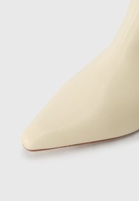 Chaussure à talons hauts en cuir beige avec un bout pointu et une texture lisse, présentant un talon fin et un design minimaliste.