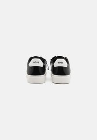 Paire de baskets basses noires et blanches avec semelles blanches, vues de dos, affichant le logo "BOSS" sur les languettes de talon.