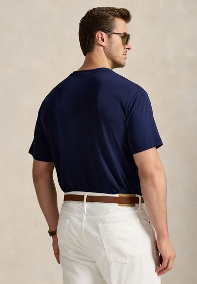 Bărbat purtând o cămașă albastru închis cu mâneci scurte, pantaloni albi cu o curea maro și ochelari de soare, cu fața spre spate pe un fundal neutru.