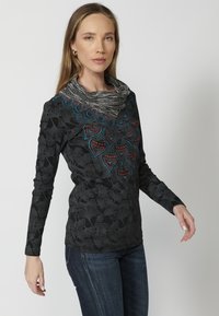 Langärmliges schwarzes Oberteil mit floralen Mustern, ausgestattet mit einem mehrfarbigen gestickten Ausschnitt und einem strukturierten Kragen. Getragen mit Jeans.