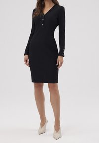 Femme portant une robe noire cintrée arrivant aux genoux avec des manches longues, des boutons argentés aux poignets et au niveau de l'encolure, associée à des talons blancs pointus.