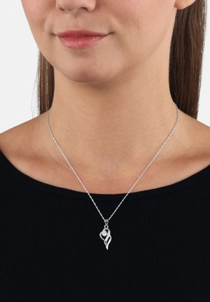 Collana con pendente a forma di cuore in argento con piccoli cristalli indossata intorno al collo di una donna sopra un top nero a costine.