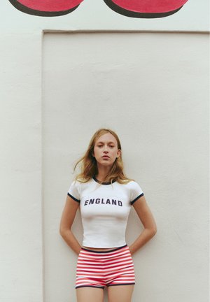Jeune femme aux longs cheveux blonds se tenant contre un mur blanc, portant un t-shirt blanc England et un short rayé rouge et blanc.