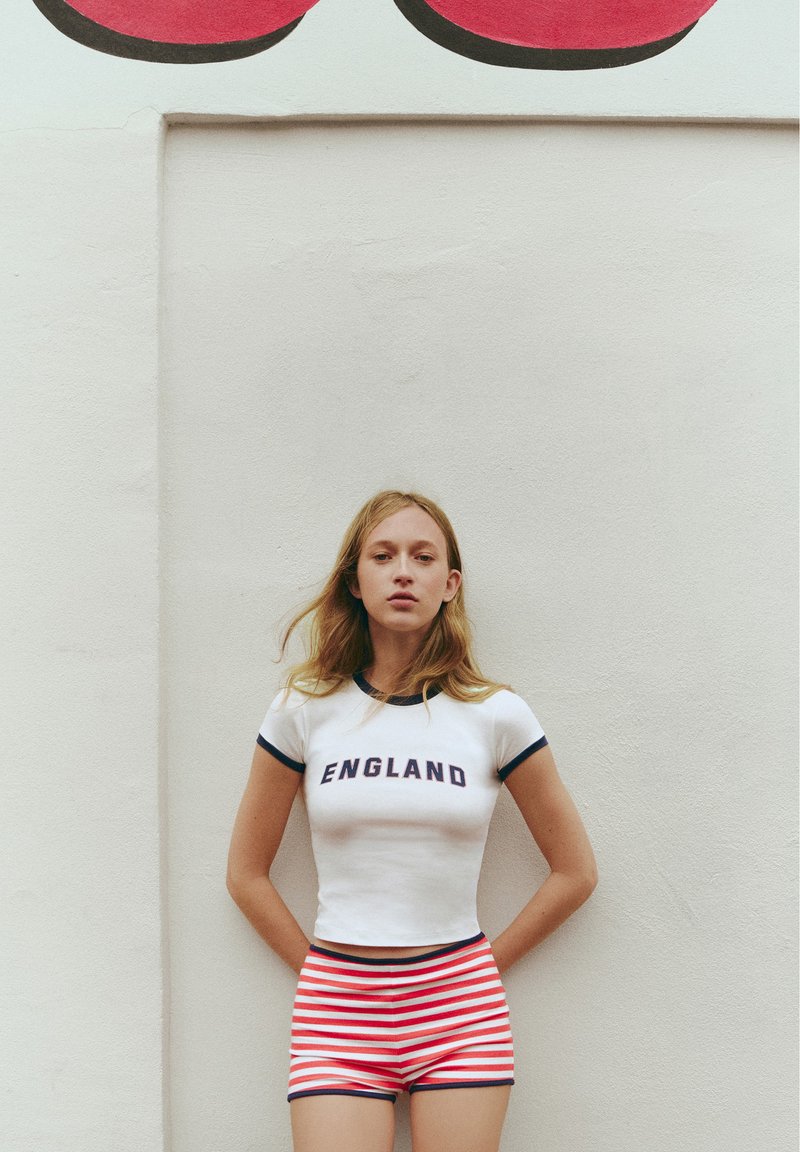 Jeune femme aux longs cheveux blonds se tenant contre un mur blanc, portant un t-shirt blanc England et un short rayé rouge et blanc.