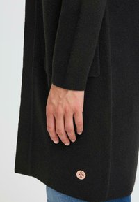 Ce cardigan en maille noire présente une finition texturée, des manches longues et un ourlet inférieur arrondi ; il inclut un petit accent logo près de l'ourlet.