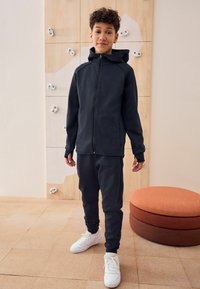 Donkergrijze zip-up hoodie met een capuchon, gecombineerd met bijpassende tapered sweatpants. Witte sneakers. Glad materiaal, minimalistisch ontwerp en een aansluitende snit.