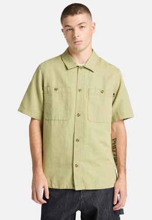 Joven con cabello corto que lleva una camisa de manga corta de color verde claro con botones y bolsillos delanteros, y jeans oscuros, de pie contra un fondo blanco.