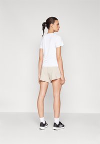 Camiseta blanca de manga corta combinada con pantalones cortos beige, mostrando un ajuste relajado. El modelo lleva zapatillas negras con acentos blancos y calcetines tobilleros.