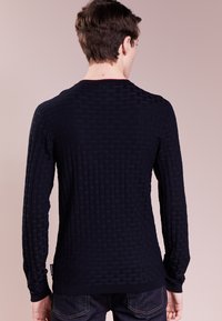 Homme vu de dos portant un pull noir à manches longues texturé et un jean sombre, sur un fond beige uni.