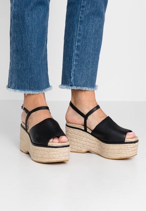 Czarne skórzane sandały na platformie z odkrytymi palcami, plecioną podeszwą z juty i regulowanym paskiem na kostkę, zestawione z dżinsami o postrzępionych nogawkach.