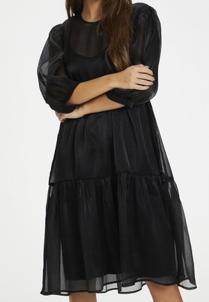 Woman wearing a black sheer layered dress with puffed sleeves, arms crossed, against a plain white background.