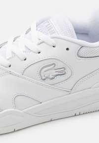 Fehér bőr sneaker, hálós részletekkel. Lapított fűzők, dombornyomott logó és texturált oldalpanelek jellemzik. Gumitalp, minimális dizájn.
