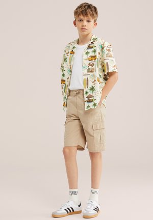Junge trägt beige Cargo-Shorts, weißes T-Shirt, offenes tropisch gemustertes Hemd, weiße Socken und weiße Sneaker mit schwarzen Streifen, steht mit Händen in den Taschen.