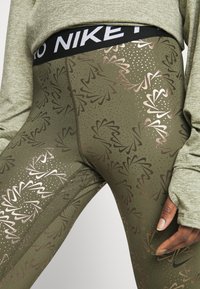 Olive zielone legginsy z metalicznymi różowymi złotymi wzorami kwiatowymi i kropeczkami, z czarnym pasem w talii z logo „NIKE”, gładka tekstura, dopasowany krój.